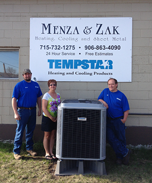 Two men and a woman standing around an air conditioning unit in front of a sign that reads. "Menza & Zak"